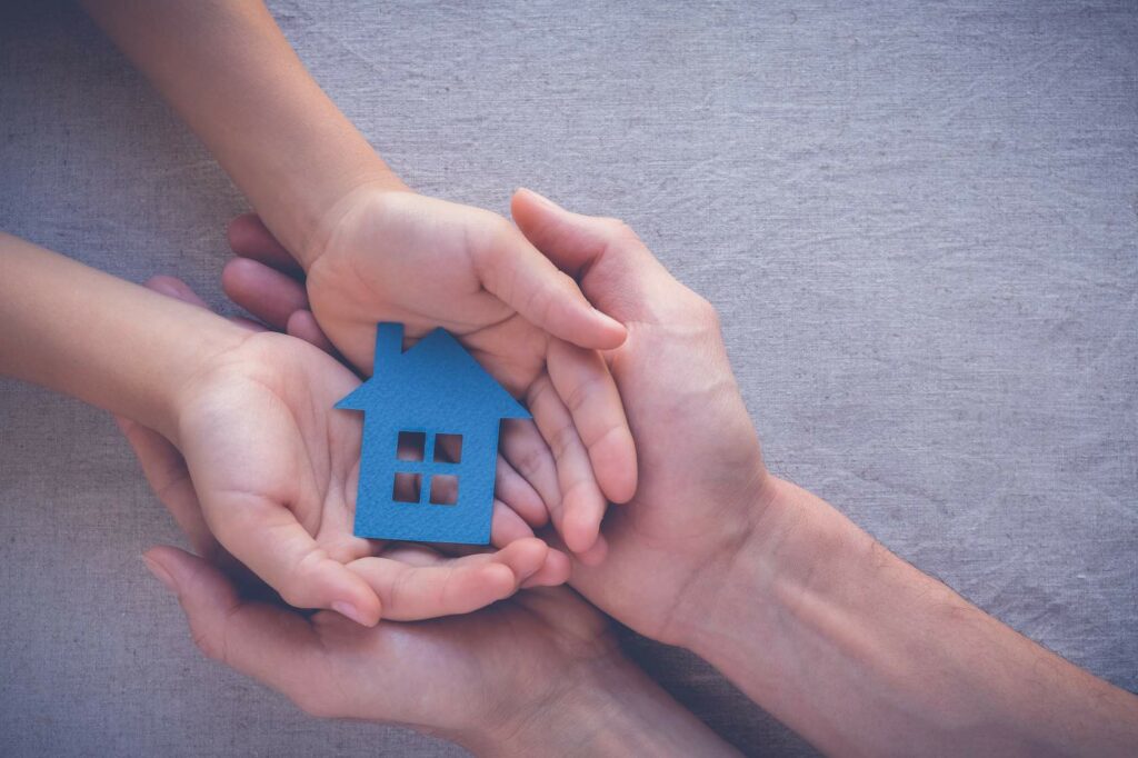 A cut out blue house being held by a child's hands supported by bigger adult hands.